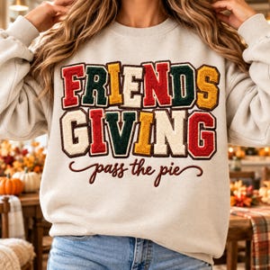 May include: A beige crewneck sweatshirt featuring the text "FRIENDS GIVING" in large, multi-colored chenille varsity-style letters with dark brown outlines, and the cursive text "pass the pie" in dark brown below the main graphic.