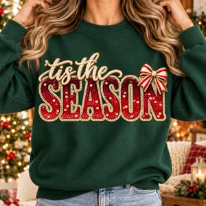 Puede incluir: Una sudadera de manga larga de color verde oscuro con el texto 'tis the SEASON' en una fuente roja de lentejuelas con borde dorado. Un lazo a rayas rojas y blancas está sujeto en la parte superior derecha de la palabra SEASON.