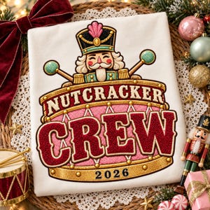 May include: A white t-shirt featuring an embroidered design of a nutcracker character emerging from a pink and gold drum. The text reads NUTCRACKER CREW 2026 in bold, textured lettering. The design includes two drumsticks and a pink plume.