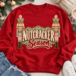 May include: A red crewneck sweatshirt featuring a graphic print of two nutcracker figures in green and pink uniforms flanking the text "NUTCRACKER Season" in a stylized font. The design is centered on the chest of the long-sleeved garment.