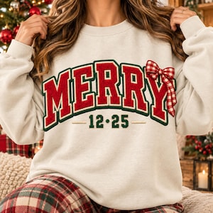 May include: A cream-colored crewneck sweatshirt featuring the word MERRY in large red letters with dark green outlines. A red and white checkered bow is attached to the letter Y. Below the word, the text 12-25 is printed in dark green.
