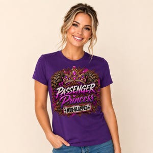 Op de afbeelding: Een paars t-shirt met korte mouwen en een grafische print van een kroon met juwelen, bandensporen en de tekst "PASSENGER Princess" in roze en witte letters, met een rechthoekig "MUD-SLAPPED"-bordje aan de onderkant en kleine roze hartjes.