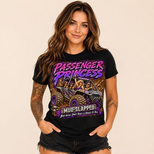 Może przedstawiać: Czarny t-shirt z nadrukiem graficznym przedstawiającym pojazd terenowy z czterema osobami w środku. Tekst brzmi "PASSENGER PRINCESS" w kolorze różowym i fioletowym, "MUD-SLAPPED" na metalowej tabliczce oraz "BAD IDEAS, DIRTY TRAILS & STORIES TO TELL" poniżej.