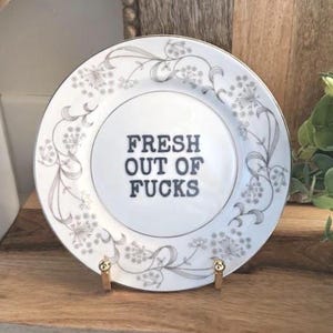 May include: A white ceramic plate featuring a silver floral pattern along the rim and the black text "FRESH OUT OF FUCKS" printed in the centre. The plate is displayed on a small gold-toned metal stand, highlighting the contrast between the delicate silver detailing and the bold, dark lettering.