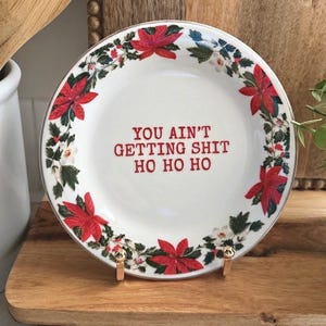 May include: A round white ceramic plate featuring a decorative border of red poinsettia flowers and green leaves. The centre of the plate displays the red text: YOU AIN'T GETTING SHIT HO HO HO. The plate is supported by a small gold-toned metal stand.