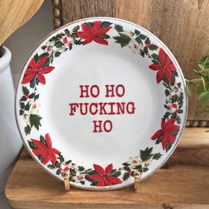 May include: A white ceramic plate with a silver rim featuring a red poinsettia and green leaf border pattern. The centre of the plate displays the red text "HO HO FUCKING HO" in a bold, sans-serif font. The plate is held upright by a gold metal stand.