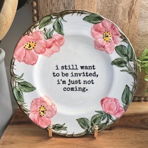 May include: A white ceramic plate with a scalloped edge featuring hand-painted pink flowers and green leaves. The centre of the plate displays the black printed text: i still want to be invited, i’m just not coming. The plate has a gold-toned rim.