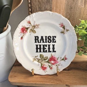 May include: A white ceramic plate with a scalloped edge and gold trim, featuring pink rose floral patterns and the black text RAISE HELL printed in the centre. The plate is displayed on a small gold metal stand.
