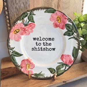 Puede incluir: Un plato redondo de cerámica blanca con un patrón floral pintado a mano de flores rosas y hojas verdes alrededor del borde festoneado. El centro del plato muestra el texto negro "welcome to the shitshow" en una fuente sans-serif simple y negrita.