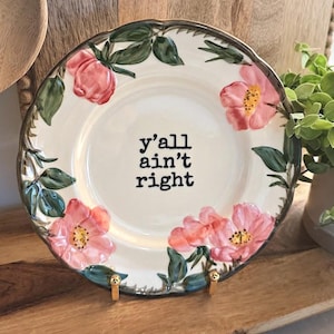 May include: A round ceramic plate with a cream-coloured centre featuring the black text 'y'all ain't right'. The rim is decorated with hand-painted pink flowers and green leaves, finished with a dark scalloped edge and supported by a gold stand.