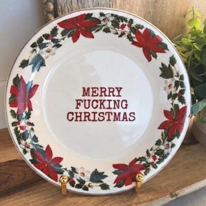 Puede incluir: Un plato de cerámica blanca con un patrón de borde de flores de pascua rojas y hojas de acebo verdes. El centro del plato muestra el texto MERRY FUCKING CHRISTMAS en letras mayúsculas rojas. El plato tiene un borde fino de color dorado.