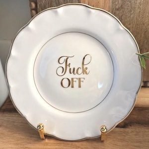 May include: A white ceramic plate with a scalloped edge and a gold-coloured rim. The centre of the plate features the text "Fuck OFF" written in a metallic gold script and block font. The plate is displayed on a small gold-coloured metal stand.