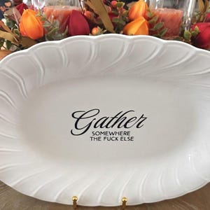 May include: An oval white ceramic serving platter with a scalloped, embossed edge. The center features black text that reads Gather SOMEWHERE THE FUCK ELSE in a combination of elegant script and bold, clean sans-serif typography.