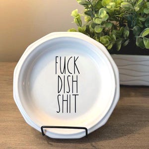 May include: A white ceramic dish with a scalloped edge and a black sans-serif font printed in the centre. The text reads FUCK DISH SHIT in three vertical lines. The dish is held upright by a thin black metal stand.