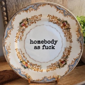 May include: A white ceramic plate featuring a decorative gold and light blue floral border with intricate scrollwork. The centre of the plate displays the black text "homebody as fuck" in a typewriter-style font, centred on the smooth surface.