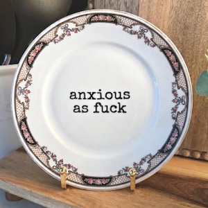 May include: A white ceramic plate with a black and pink floral border pattern and a lattice design. The center of the plate features the black text "anxious as fuck" in a typewriter-style font. The plate is held upright by a gold metal stand.