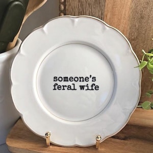 May include: A white ceramic plate with a scalloped edge and a thin gold rim. The centre of the plate features the black text 'someone's feral wife' in a typewriter-style font. The plate is displayed on a small gold-toned metal stand.