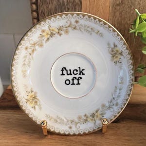 May include: A white ceramic saucer featuring a delicate floral pattern in shades of yellow and green along the scalloped rim, with the text "fuck off" printed in bold black typewriter-style font in the centre of the circular indentation.