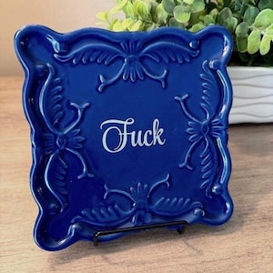 May include: A square, deep blue ceramic decorative plate featuring an ornate, raised relief border with scrolling patterns. The word "Fuck" is printed in white, cursive script in the center of the plate. The plate is displayed on a black metal stand.