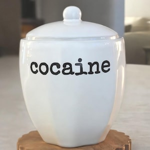 May include: A white ceramic canister with a matching lid and a rounded knob handle. The word "cocaine" is printed in black, lowercase, sans-serif lettering on the front of the smooth, glossy, slightly faceted container body.
