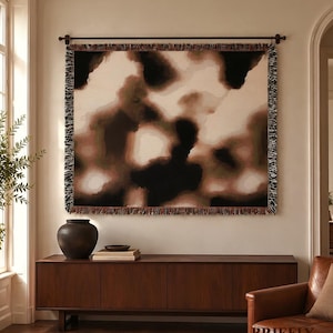 May include: A rectangular woven wall tapestry featuring an abstract pattern of blended black, dark brown, and cream tones. The textile has a fringed border on all four sides and is suspended from a dark metal rod mounted on the wall.