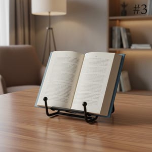 May include: A black metal book stand holding an open hardcover book with white pages and black text. The stand features two curved arms with rounded tips that support the bottom edges of the book, keeping it open for reading on a flat surface.
