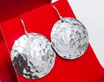 Puede incluir: Un par de pendientes colgantes redondos de tono plateado con una textura de superficie martillada y reflectante. Los pendientes cuelgan de finos ganchos de alambre curvados y se muestran sobre un joyero de color rojo brillante.