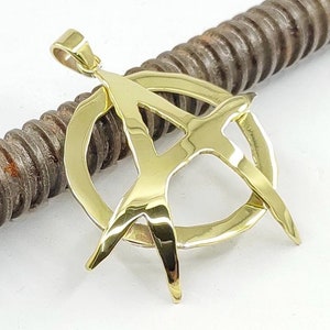 May include: A gold pendant necklace featuring an anarchy symbol. The pendant is a circle with a stylized letter A inside.