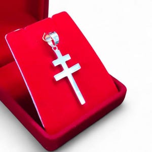 Pode incluir: Um pingente de cruz de barra dupla em tom prateado com acabamento metálico polido e uma pequena argola circular no topo, apresentado sobre um inserto de veludo vermelho vibrante dentro de uma caixa de joias vermelha a combinar.