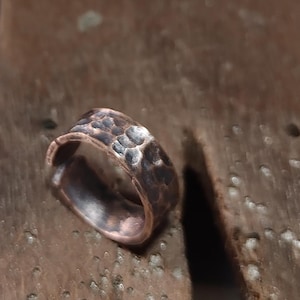 May include: A hammered copper ring with a textured surface. The ring has a dark, mottled appearance with a band that is approximately 2 cm wide. The metal has a rustic, aged look.