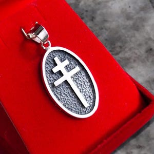 May include: Silver pendant with a double-barred cross design inside an oval frame. The pendant has a textured background and a polished silver bail. The pendant is displayed on a red surface.