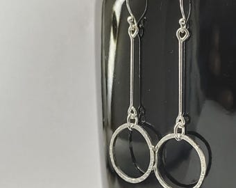 May include: A pair of silver-toned drop earrings featuring a long, slender vertical bar attached to a circular hoop at the bottom. The hoops have a textured, hammered finish, and the earrings are suspended from simple curved ear wires for hanging.