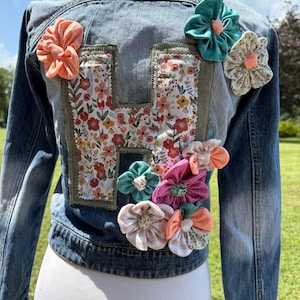 May include: A blue denim jacket featuring a large letter H appliqued on the back. The letter is filled with a white fabric printed with small pink, orange, and yellow flowers. Several fabric flowers in shades of peach, teal, and white are attached.