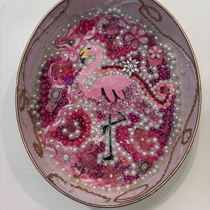 May include: An oval-shaped decorative container filled with a collage of pink and white beads, pearls, and floral embellishments. A central pink flamingo figure with a black beak and legs is surrounded by various textures and shiny accents.