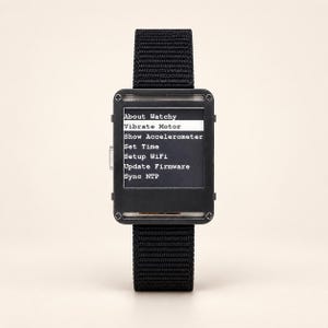 May include: A black rectangular digital watch with a woven nylon strap. The screen displays a menu with the text: About Watchy, Vibrate Motor, Show Accelerometer, Set Time, Setup WiFi, Update Firmware, and Sync NTP, with Vibrate Motor highlighted.