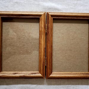 May include: A hinged double picture frame constructed from medium-toned wood with a visible grain pattern. The two rectangular frames are connected by metal hinges, each featuring a plain brown backing board within the wooden borders.