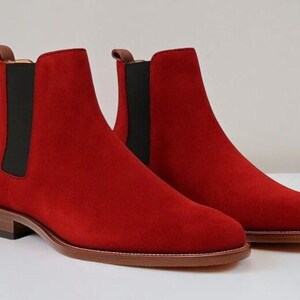 May include: A pair of vibrant red suede Chelsea boots featuring black elastic side panels, a brown leather heel, a matching brown leather sole, and a small brown pull tab at the top of the ankle collar for a sleek and functional footwear design.