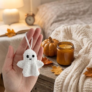 May include: A small white crocheted ghost ornament featuring two black embroidered eyes and a tiny black mouth. The ghost has a rounded head, outstretched arms, and a flared base, with a white yarn loop attached to the top for hanging purposes.