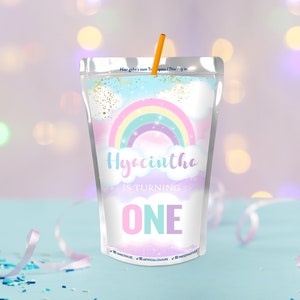 Op de afbeelding: Een kleurrijke regenboog-thema sapzak met de tekst "Hyacintha wordt één" en een roze, blauwe en paarse "EEN" op een witte achtergrond. De zak is versierd met gouden glitter en wolken. De tekst "Geen zoetstoffen, geen kunstmatige kleurstoffen, geen conserveermiddelen" staat onderaan de zak.