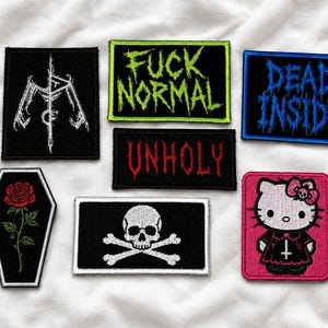 Puede incluir: Una colección de siete parches bordados con varios diseños: un sigilo blanco sobre negro, texto verde neón FUCK NORMAL, texto azul DEAD INSIDE, texto rojo UNHOLY, una rosa roja en un ataúd, una calavera con huesos cruzados y un gato rosa.
