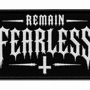 May include: A rectangular black embroidered patch featuring the white text "REMAIN FEARLESS" in a stylized, sharp-edged font. The design includes a small white cross symbol positioned below the word "FEARLESS" and a black stitched border.
