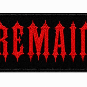 May include: A rectangular embroidered patch with a black background and the word REMAIN stitched in a bold, red, gothic-style font. The lettering features sharp, pointed edges and a textured thread finish, with a black stitched border surrounding the entire piece.