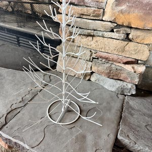 May include: A white metal display stand shaped like a leafless tree with a central vertical pole and multiple branching arms extending outward. The structure features a circular base and is designed with numerous thin prongs for hanging items.