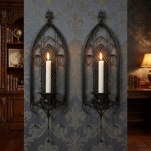 May include: A pair of black metal wall sconces featuring an arched, gothic-inspired frame with a lattice back panel. Each sconce holds a single white taper candle in a central cup, with a decorative finial at the top and hanging bead chains.