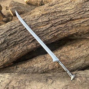 Könnte beinhalten: Ein silberfarbenes Schwert mit einer geschwungenen, spitzen Klinge, die entlang der Mitte komplizierte, geätzte Linienmuster aufweist. Der Griff ist metallisch mit detaillierten rankenartigen Gravuren und einem ausgestellten Knauf.