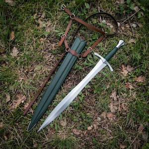 Pode incluir: Uma espada de aço com punho verde-escuro e uma bainha verde-escura a condizer. A bainha possui um arnês de couro castanho com fivelas e anéis de metal para transporte. A espada tem uma guarda prateada e uma lâmina pontiaguda com nervura.