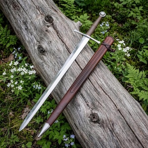 May include: A double-edged steel sword with a straight crossguard and a brown leather-wrapped grip, accompanied by a matching brown leather scabbard featuring a silver-toned metal chape at the pointed end, resting on a weathered wooden log.