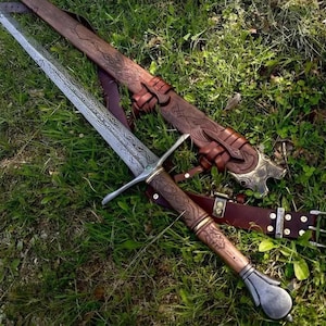 Può includere: Una spada in acciaio con lama incisa, guardia a croce e impugnatura in legno marrone con intagli complessi. La spada è accompagnata da un fodero in pelle marrone abbinato con motivi in rilievo e una cintura in pelle con fibbie metalliche.
