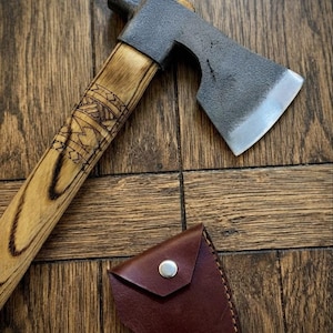 Può includere: Un'accetta con una testa in metallo scuro e testurizzato e un manico in legno chiaro caratterizzato da intricati motivi geometrici scuri pirografati. È incluso un piccolo fodero in pelle marrone separato con un bottone a pressione in metallo argentato.