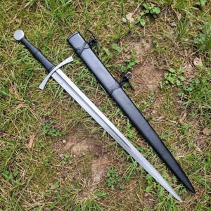 Op de afbeelding: Een stalen zwaard met een lang, tweesnijdend lemmet, een rechte pareerstang en een zwarte greep met een ronde pommel. Naast het zwaard ligt een bijpassende zwarte schede met lederen koorden voor bevestiging.